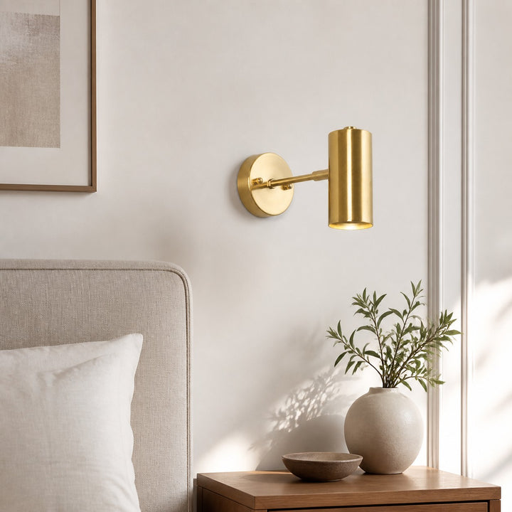 Brass cylinder wall sconce beside a neutral upholstered bed